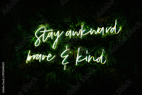 A fun neon sign glowing among greenery that reads, stay awkward, brave and kind