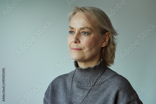 Natural light portrait of a mature woman with short blonde hair and blue eyes, wearing a gray turtleneck sweater, looking thoughtfully to the side against a minimal neutral background.