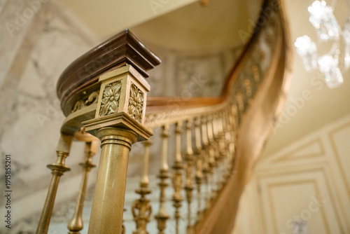 A close-up of an ornate golden stair railing features a floral carving and polished wooden handrail. The elegant curved staircase in the background glows under a crystal chandelier.