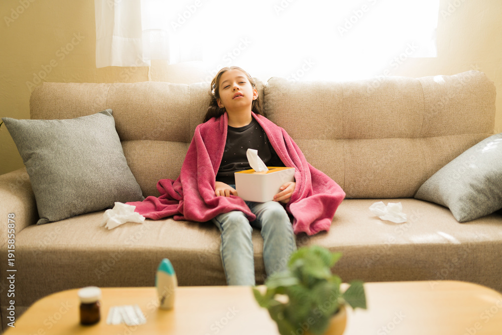 Fototapeta premium Tween girl feeling sick at home with flu symptoms, covered in blanket and holding tissues