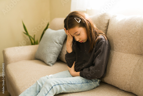 Obraz na płótnie Young child experiencing severe pain, holding her head and stomach while sitting on a couch