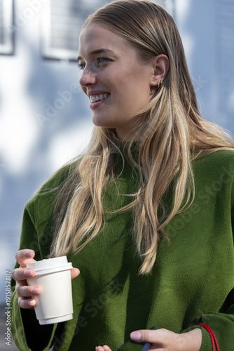Female standing outside near pale wall, smiling in green sweater, holding cup, keys, red-strap