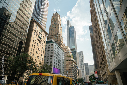 A yellow taxi drives down a bustling street in Midtown Manhattan. Historic and modern skyscrapers rise into a bright summer sky.