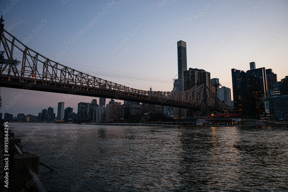 Fototapeta premium The Queensboro Bridge stretches across the East River as the last light of day fades over Manhattan's skyline. Reflections shimmer on the calm water, and the city prepares for nightfall.