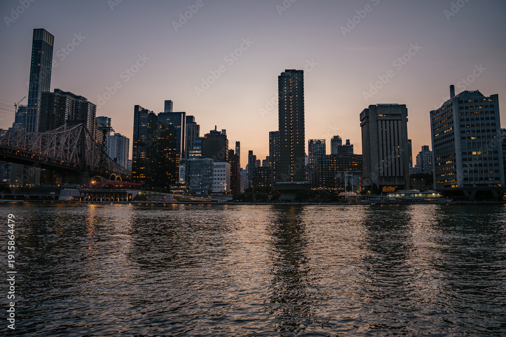 Naklejka premium The Manhattan skyline is seen across the East River during golden hour, with high-rise buildings glowing in the fading light. The water reflects the warm tones of the sunset and the city's illuminated