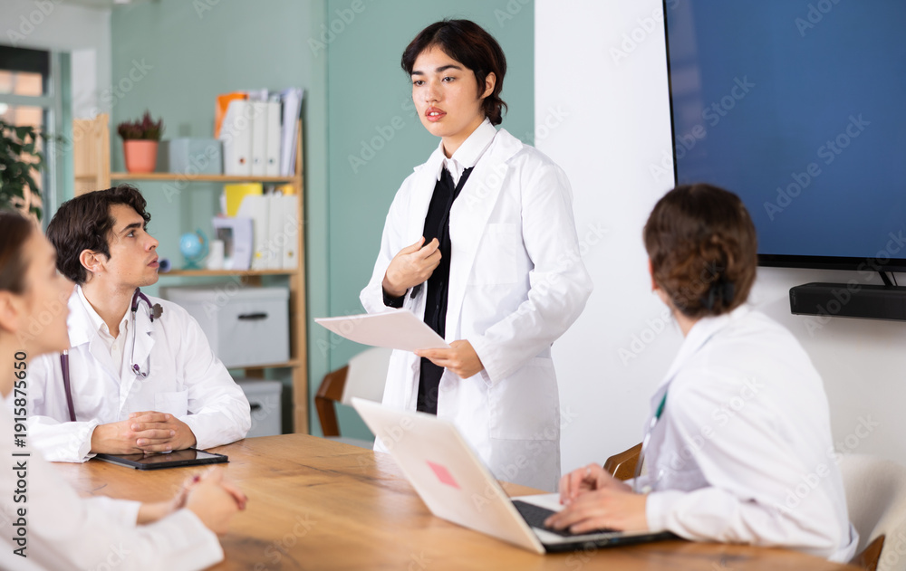 Fototapeta premium Young female doctor in medical gown holds meeting at table with group of colleagues