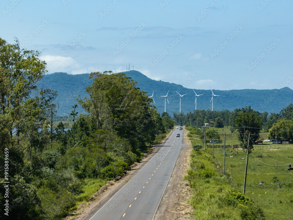 Obraz premium Rural road leading to wind turbines with mountain landscape under blue sky