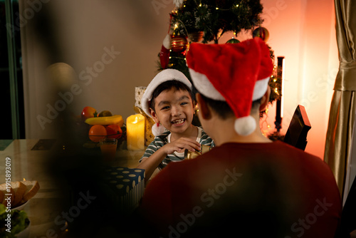 Wallpaper Mural Father and son wearing Santa hats celebrating Christmas at home, smiling at dinner table with decorated tree, wrapped gift and candlelight, warm family holiday tradition, festive evening Torontodigital.ca