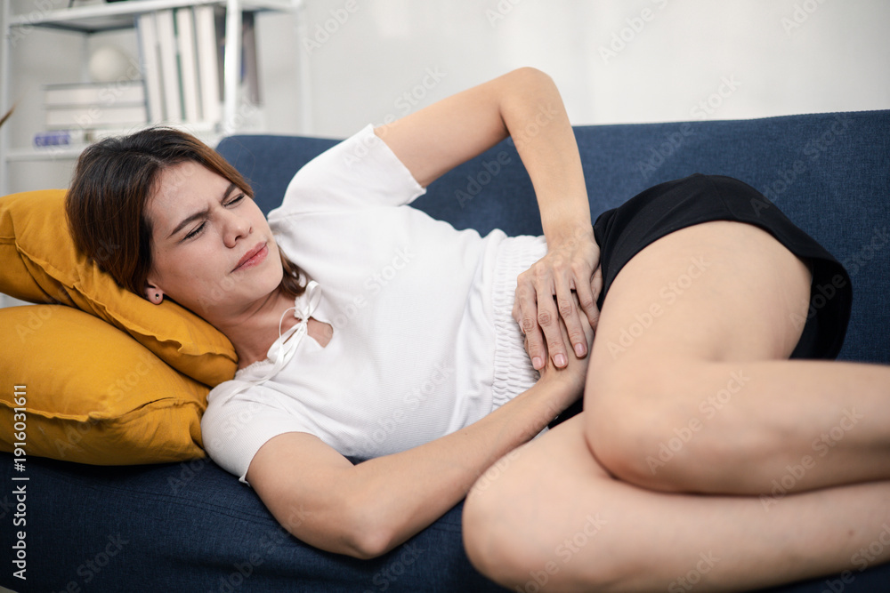 Obraz premium Woman sitting on sofa holding her stomach in discomfort, depicting abdominal pain, menstrual cramps, or digestive issues for health and medical content use.