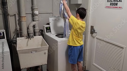 Young man takes clean wet laundry out of a washing machine and shakes it to loosen the fabric before drying. The action highlights routine laundry care, clothing handling, and everyday household work.