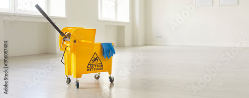Φωτογραφία Horizontal photo, banner view of yellow mop bucket with caution sign stands inside empty, spacious room with shiny floors and panoramic windows