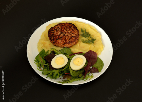 Plate of delicious lunch, fried meat patty with dill sprig on mashed potatoes and mix of culinary herbs with sliced ​​boiled egg on black background.