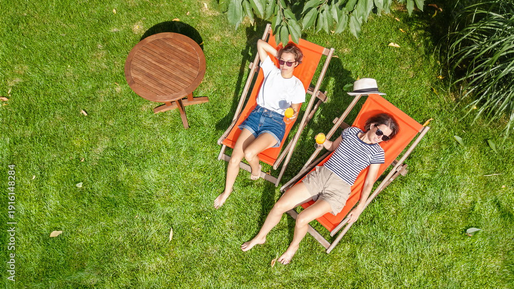 © Iuliia Sokolovska - Young girls relax in summer garden in sunbed deckchairs on grass, women friends have fun outdoors in green park on weekend, aerial drone view from above