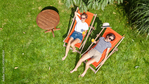 Young girls relax in summer garden in sunbed deckchairs on grass, women friends have fun outdoors in green park on weekend, aerial drone view from above