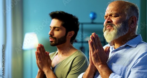 Indian senior father and young son praying with folded hands and chanting mantra as daily devotional ritual practice in modern Hindu home living room, expressing faith, spirituality and family unity