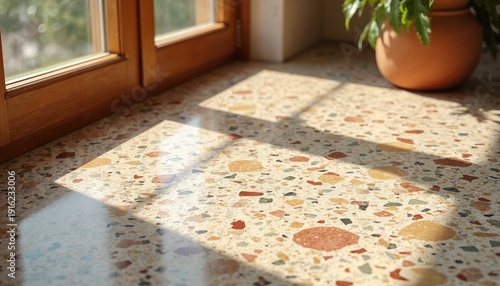 Earthy toned terrazzo floor in sunlit room. Colorful stone chips form speckled pattern on beige surface. Wooden window frame casts light shadows. Indoor plant sits nearby.