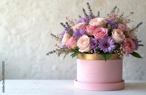 Pink round flower box with roses and purple flowers sits on white table. Floral arrangement with pastel blooms on textured wall background. Perfect gift for special occasions and holidays.