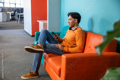 Εκτύπωση καμβά Asian man holding smartphone and wearing headphones, sitting on orange couch in
