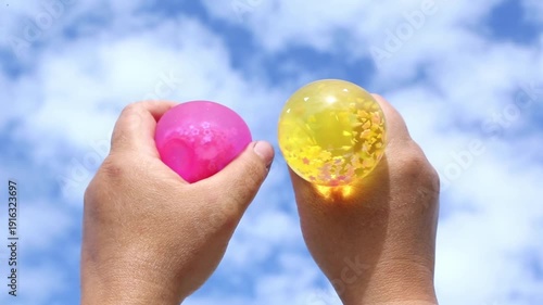 Close-up of two hands squeezing colorful gel stress balls against a bright blue sky. Fingers press, release, rotate the toys as shapes deform and recover, showing elastic motion.