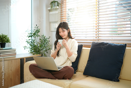 Free Download A woman smiles while sitting and relaxing on a couch in a living room, holding a mug and using laptop - GFXLoad