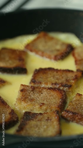 Delicious sweet french toast bread pudding cooking in an egg mixture inside a hot black frying pan, a simple and tasty homemade breakfast dish representing a comforting culinary process. Vertical