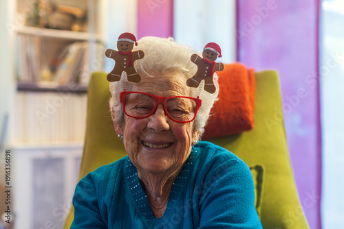 Festive portrait of smiling senior woman wearing Christmas gingerbread glasses and santa hat boppers at home. Cheerful elderly grandmother celebrating the holidays. Authenti