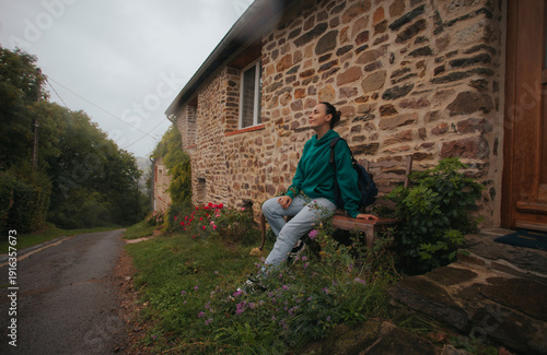 Young woman sitting on a wooden bench outside a rustic stone house by a quiet village road, peaceful countryside lifestyle and travel concept.