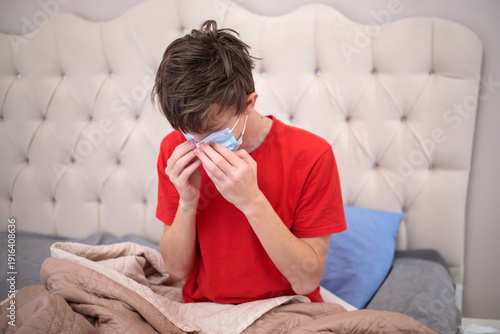 Fotografia Sick teenage boy wearing medical mask and covering nose, suffering from flu or c