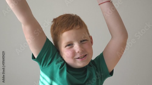 Adorable handsome boy raising hands and dancing rejoicing achievement. Happy young kid looking into camera with joy emotions on his face. Portrait of child with freckles indoor. Slow motion Close up