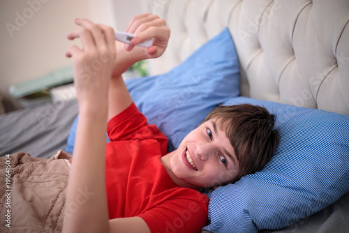 Cuadro en lienzo Recovered teenager boy smiling and looking at thermometer while lying in bed, sc