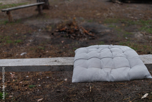 A cushion rests on a wooden bench close to a fire pit in a natural setting at dusk