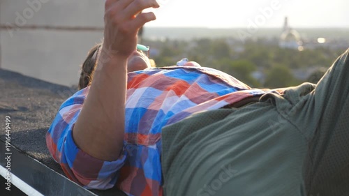 Handsome man in sunglasses lying on roof and smoking cigarette at cityscape background. Young guy relaxing and enjoying moment. Man blowing smoke outdoor. Beautiful view. Dolly shot Slow mo Close up