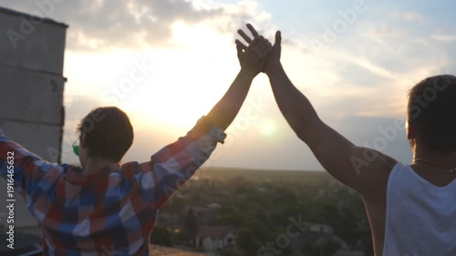 Unrecognizable gay couple standing on roof and rejoices life with cityscape at background. Friends raising hands showing joyful emotions. Men at rooftop and victoriously outstretching arms up. Slow mo