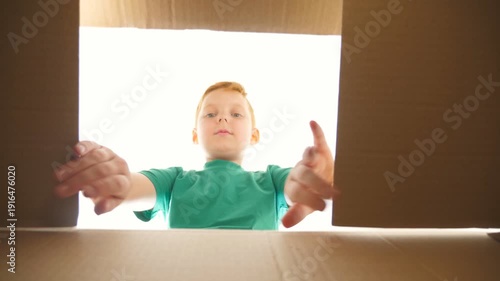 Small red-haired boy looking in parcel box with positive emotions and feelings at face. Young kid opening carton box and pulling out gift from it. Little child showing joy and surprise. Slow motion