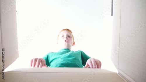 Small red-haired boy looking in parcel box with positive emotions and feelings at face. Young kid opening carton box and pulling out gift from it. Little child showing joy and surprise. Slow motion