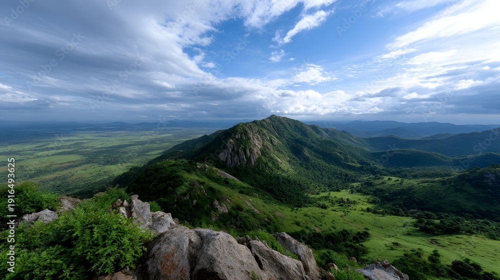 Fototapeta premium Panoramic vista from a rocky mountain summit overlooking lush green hills and a vast valley under a dramatic cloudy sky
