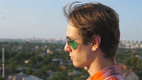 Profile of young man in sunglasses looks at cityscape from roof and enjoys beautiful view. Handsome guy watching panorama of the city from rooftop of high-rise building. Side view Close up Slow motion