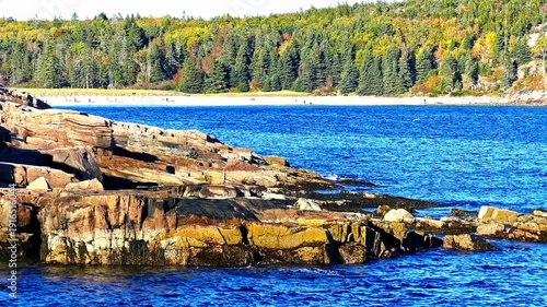Scenic view of the rugged, rocky coast of Maine with gentle waves breaking. A Sandy beach and a dense forest are visible in the distance