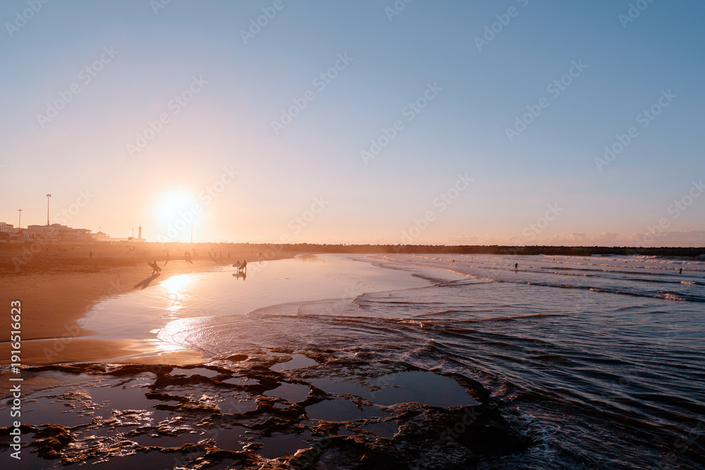 Obraz premium Peaceful scenic view of the sun setting over a calm beach, reflecting on the water with distant silhouettes of people enjoying the moment.