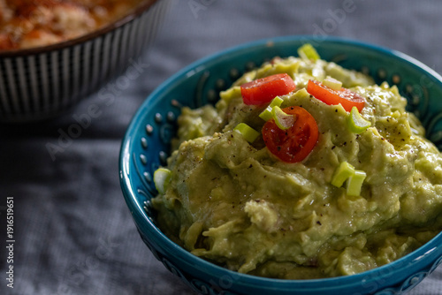 Green mashed avocado dip topped with chopped tomatoes and scallions in a blue ceramic bowl on dark fabric background