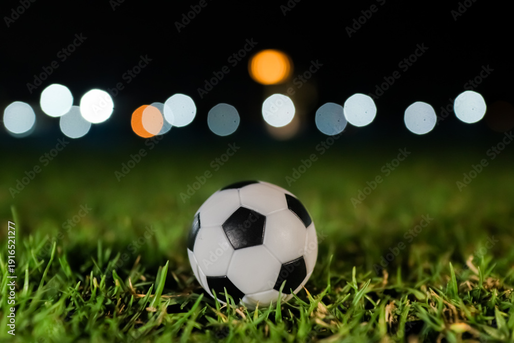 Naklejka premium Soccer ball resting on green stadium grass at night with soft glowing bokeh lights, capturing the dramatic anticipation of a global football tournament and competitive match atmosphere.