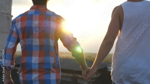 Camera follow gay couple runs on roof holding hands of each other at sunset time. Friends spending time together outdoor. Concept of male friendship. Rear back view Slow motion Close up