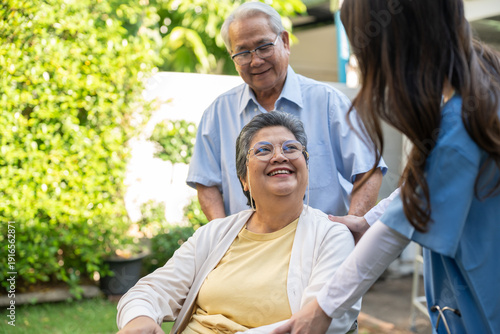 Asian nurse woman taking care of senior couple in wheelchair at garden. Happy elderly patients receiving home healthcare service professional caregiver supporting retired people lifestyle.