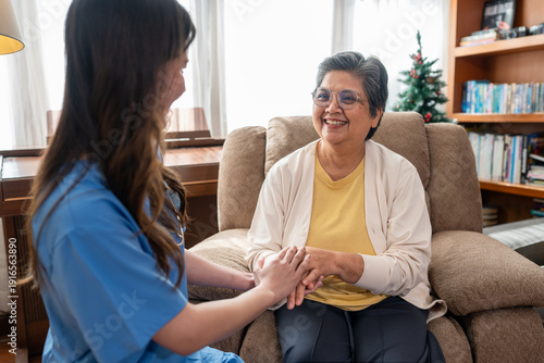 Smiling senior woman sitting on armchair holding hands with female caregiver for emotional support at home. Happy Asian elderly patient receiving professional nursing care and empathy in living room