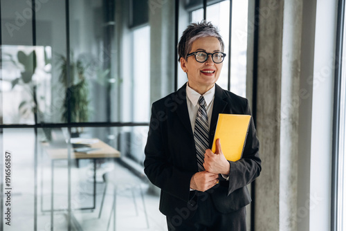 Wallpaper Mural Businesswoman in a black suit holding a yellow folder stands in a modern office with large windows and minimalist furniture, showcasing a professional environment Torontodigital.ca