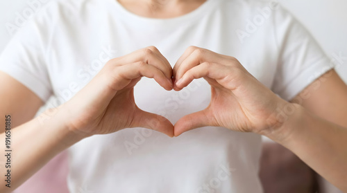 A person forming a heart shape with their hands, symbolizing love, care, and affection, with a white t-shirt and a blurred background, conveying a sense of tenderness and warmth.