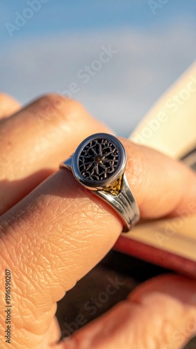 Close up of a detailed silver ring with intricate dark metalwork on a finger near an open book under bright sunlight