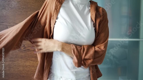 Cropped view of young woman putting on brown shirt and fastening buttons in front of wardrobe. Everyday dressing process, neat appearance and anonymous lifestyle routine.