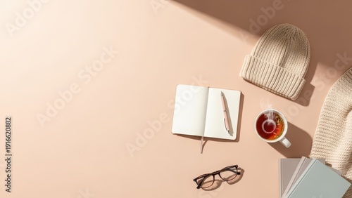 A cozy study flatlay featuring an open notebook with a pen, glasses, a warm beanie, and a cup of tea bathed in soft sunlight.