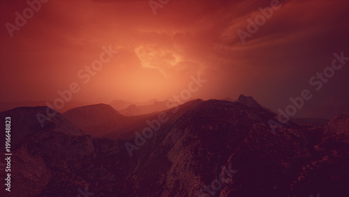 Tableau sur toile Warm light bathes the rugged mountains as the sun sets behind distant peaks, creating a serene atmosphere filled with soft hues of orange and red against the darkening sky